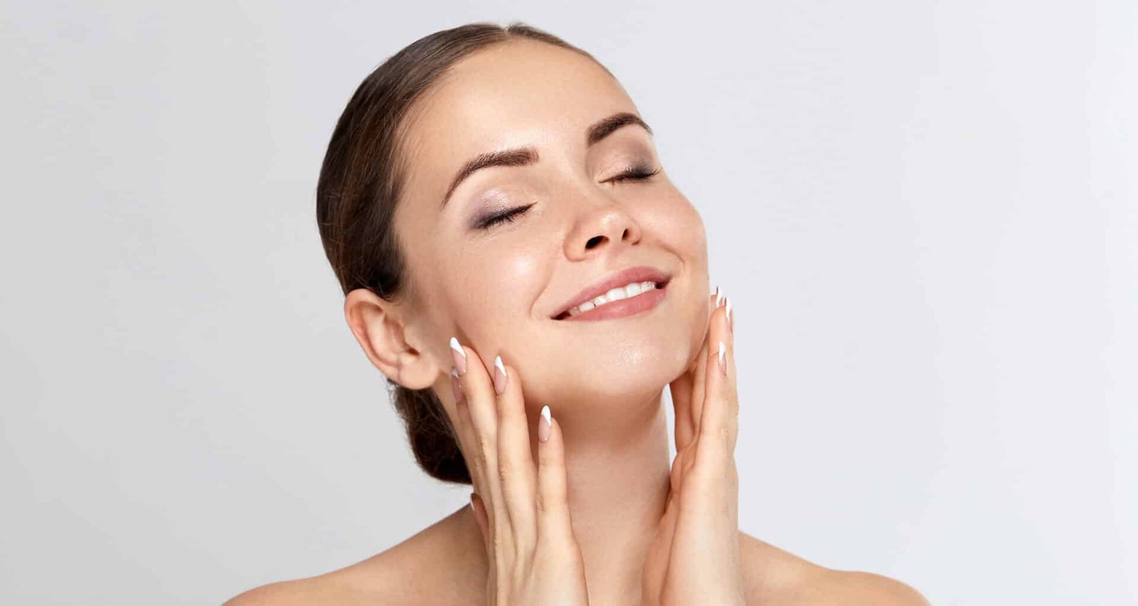 Une femme avec une peau éclatante touche son visage tout en souriant légèrement. Elle a les cheveux attachés et le teint lumineux, le fond est blanc. La photo met en avant une routine de soins de la peau.