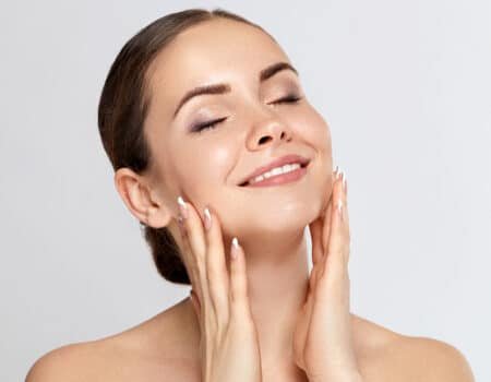 Femme souriante avec les yeux fermés, touchant son visage avec ses mains. Elle a une peau lisse et lumineuse. Son maquillage est léger, et elle porte des ongles manucurés. L'arrière-plan est clair et neutre.