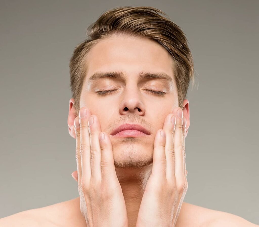 Un homme avec les yeux fermés, se touchant le visage avec les mains. Il a les cheveux bruns et mi-longs, et se tient devant un fond gris. Cette image pourrait illustrer des soins de peau ou des traitements comme l'epilation laser.