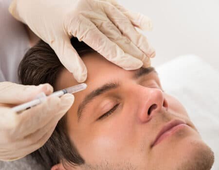 Un homme allongé sur une table de soins tandis qu'un professionnel de la santé applique un produit injectable sur son front, les mains protégées par des gants.