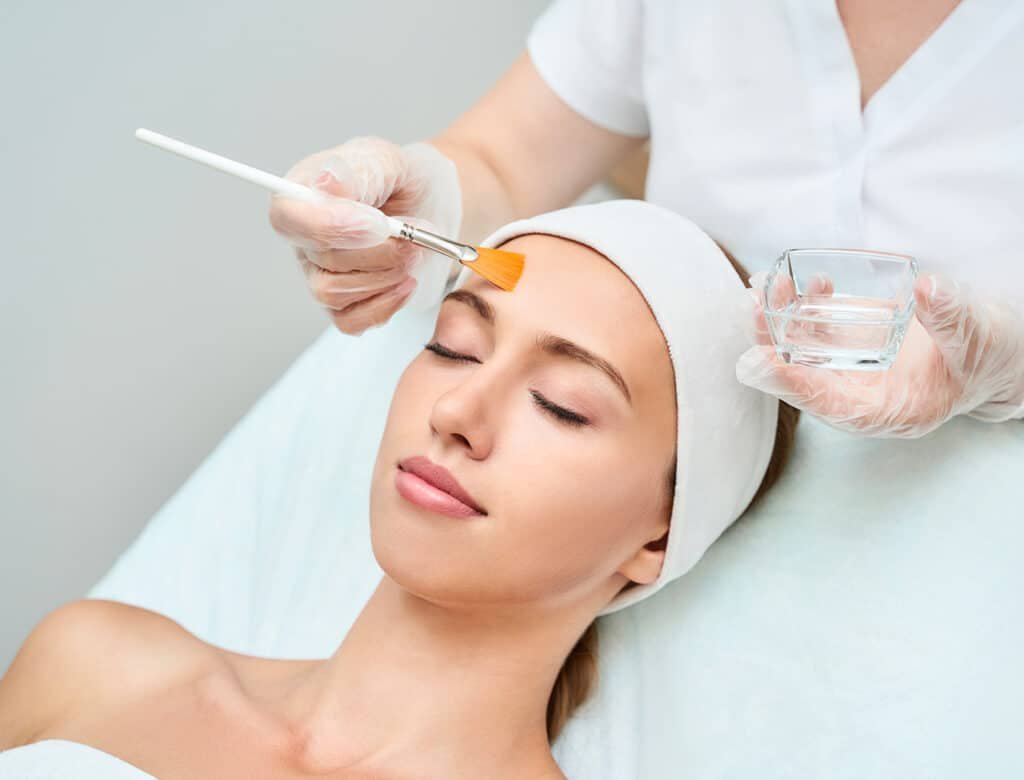 Une femme recevant un soin du visage, allongée sur une table de traitement. Une esthéticienne applique un produit sur son front avec un pinceau, tandis qu'un bol d'eau est à proximité.