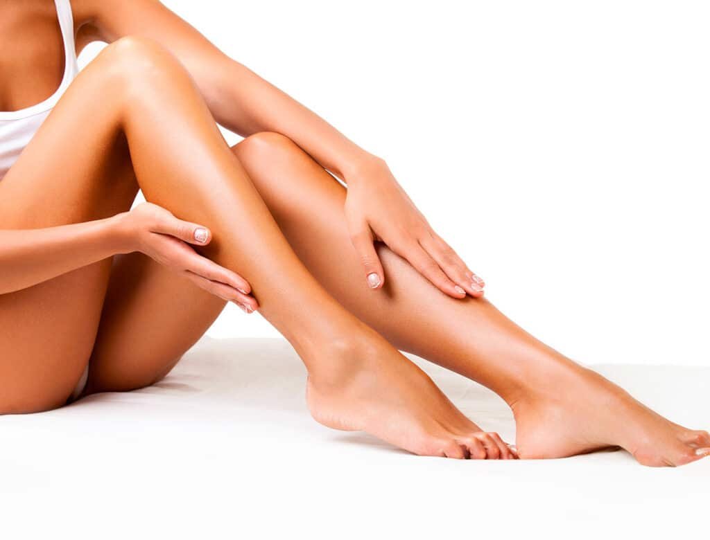 Une personne assise sur un fond blanc, tenant ses jambes. La main de gauche est posée sur la cuisse, et la main droite touche la cheville. La peau est bien hydratée et lumineuse.